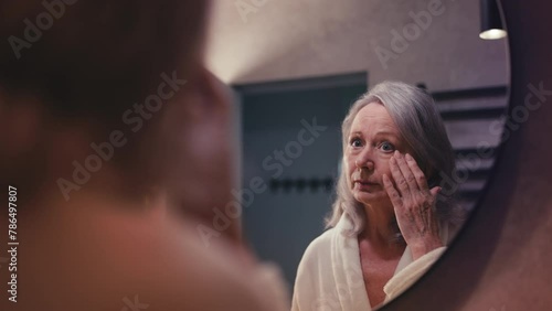 Unhappy senior lady displeased with her skin and wrinkles, looking in mirror photo