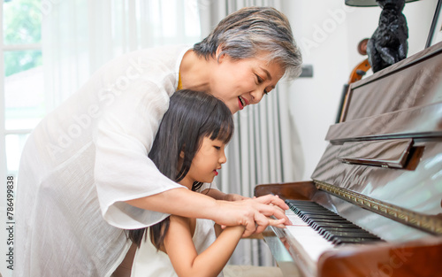 Asian elderly grandmother teaching little sweet girl playing piano, smiling with happiness, staying at cozy home, spending time together. Family, Love, Education Concept.