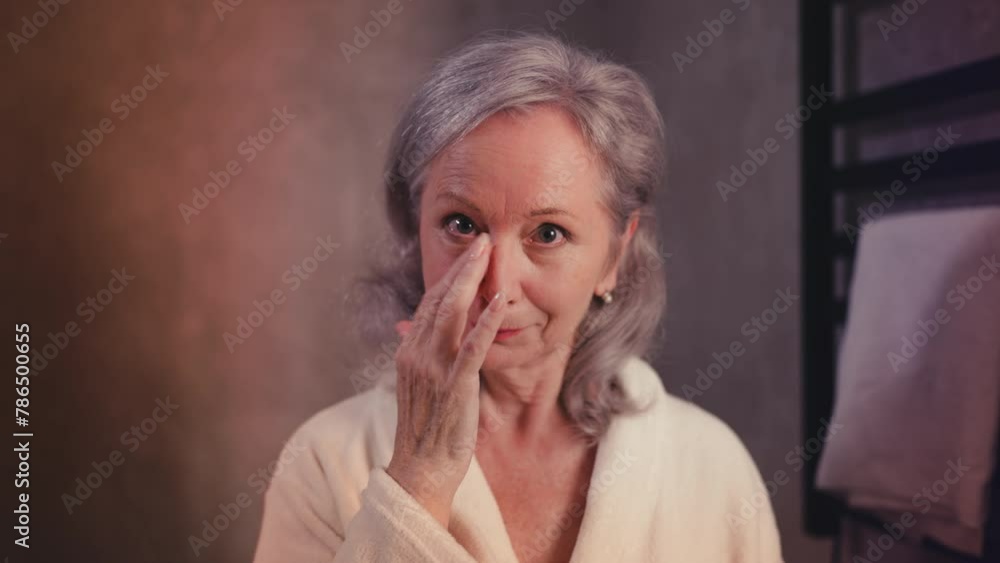 Happy senior female applying undereye cream in front of mirror, beauty rituals Happy senior female applying undereye cream in front of mirror, beauty rituals