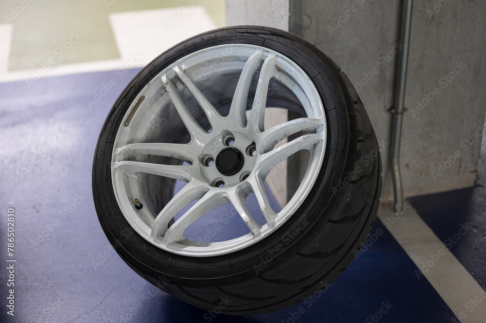 A sports car wheel resting on the pillar of a parking lot. It is a white 6-spoke wheel with a semi-slick tire.