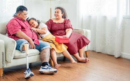 Three people asian family including happy father, husband with disable prosthetic leg, surprising little daughter girl, giving red gift box to celebrate birthday at home with love. Healthcare Concept.