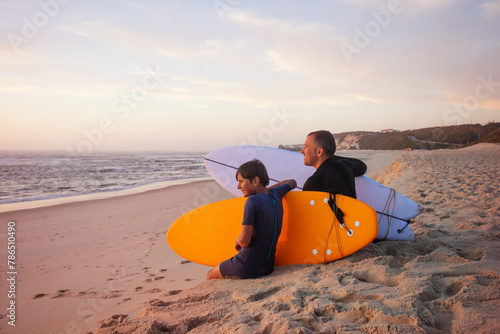 Photography A parent and his son smiling sit with their surfboards on sand