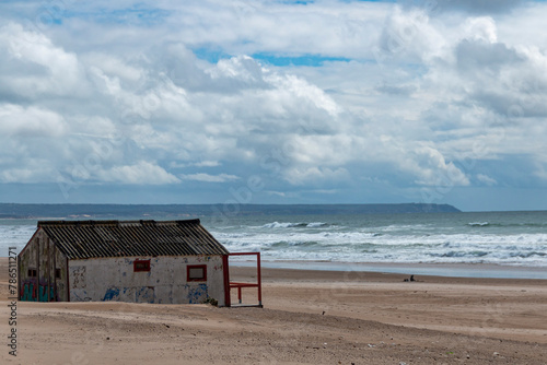 Abandoned wooden house on a beach with the sea in the background and a cloudy blue sky © SaucE ReQuEs