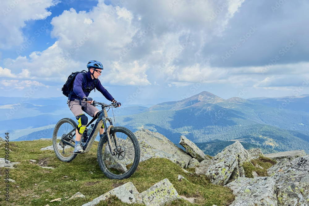 Cyclist man riding electric bike outdoors. Male tourist resting on the top of hill, enjoying beautiful mountain landscape, wearing helmet and backpack. Concept of active leisure.