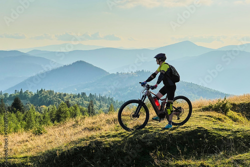 Cyclist man riding electric bike outdoors on sunny day. Male tourist resting on grassy hill, enjoying beautiful mountain landscape, wearing helmet and backpack. Concept of active leisure.