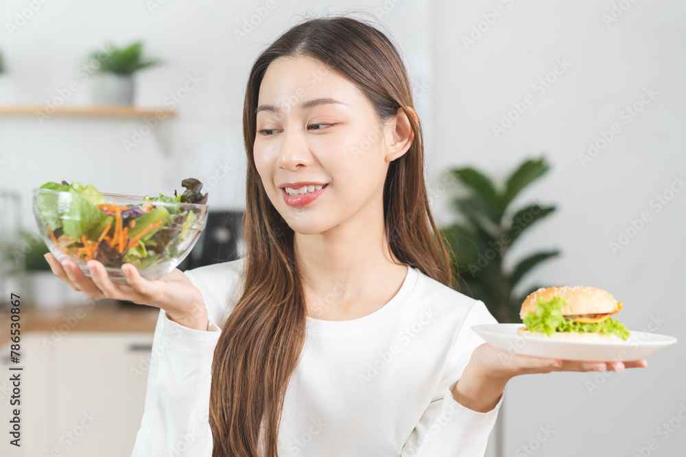 Diet, Dieting asian young woman, girl confused choose, choice eating food, hand holding bowl of vegetable salad and hamburger when hungry in kitchen at home. Temptation of female weight loss person. Diet, Dieting asian young woman, girl confused choose, choice eating food, hand holding bowl of vegetable salad and hamburger when hungry in kitchen at home. Temptation of female weight loss person.