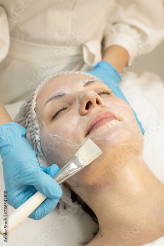 Cosmetologist in blue gloves applying poppy seeds with a brush on the patient's face, beauty clinic.