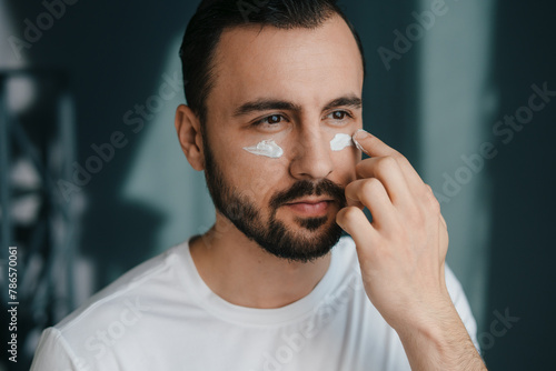 Closeup of bearded handsome man applying under eye cream at home. Good looking man using beauty products. Moisturising or nourishing skin