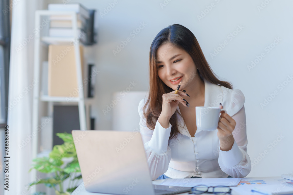 A beautiful woman is sitting at her desk and looking at her laptop. She is holding a coffee cup and smiling. A beautiful woman is sitting at her desk and looking at her laptop. She is holding a coffee cup and smiling.