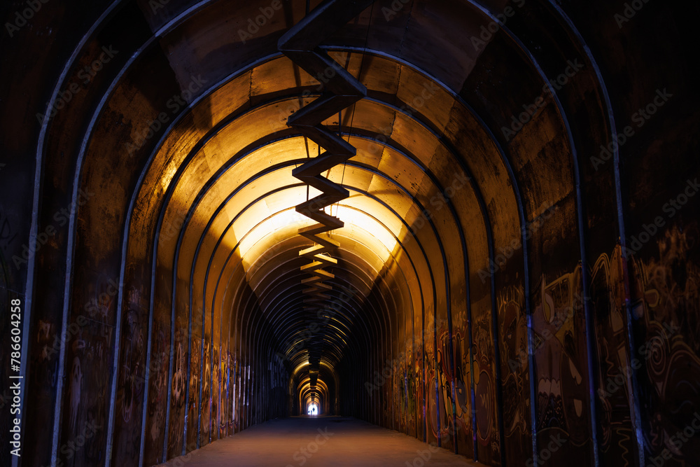 Mysterious graffiti-filled tunnel with a spiraling walkway, illuminated by warm yellow light.