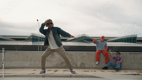 Handsome break dancer enjoy perform freestyle movement with asian friend cheering behind. Hipster listen to music from headphone and looking at camera. Street dance. Outdoor sport 2024. Sprightly.