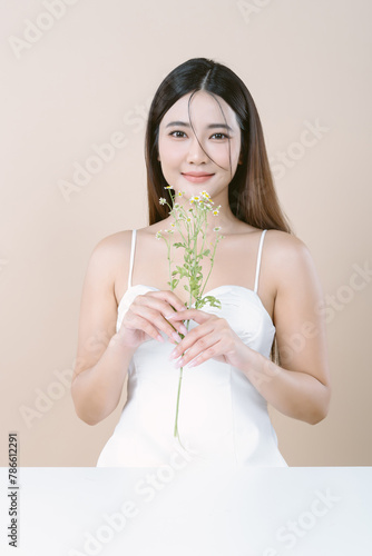 Smiling beautiful Asian woman in white dress holding Gypsophila flower and looking at camera on beige background. Skin care beauty treatments concept. White model with clean, health skin of face.