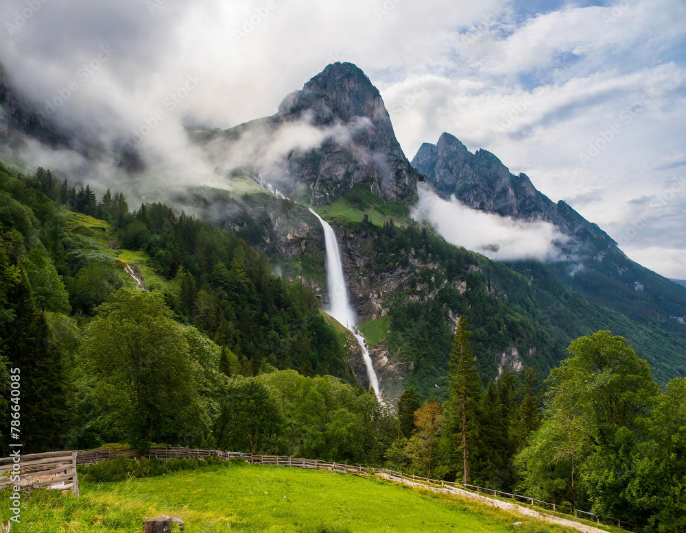 Obraz premium A mountain shrouded in mist and clouds, with a waterfall cascading from its peak, occupies the center of the photo
