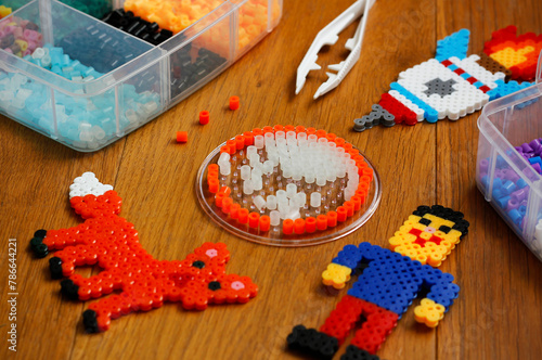 Colorful iron beads (perler beads or iron beads) in a box, art toys created from them and white tweezers on wooden background. Toy developing children's imagination. Concept of children's creativity.