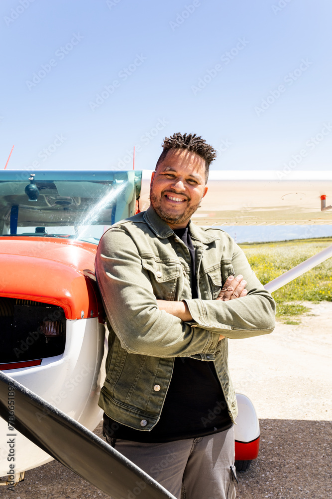 Fototapeta premium Vertical photo of a dark-skinned man poses next to a red and white plane. The Dominican adult poses with his arms crossed looking at the camera. Light aircraft concept.