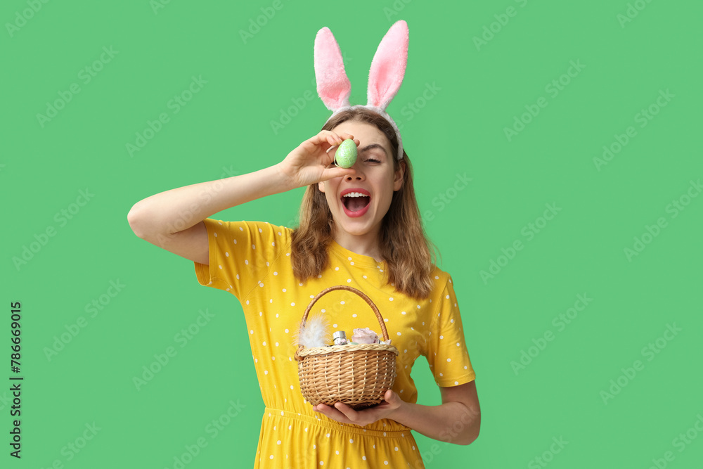 Happy smiling young woman in bunny ears headband holding basket with makeup products, flowers and Easter egg on green background