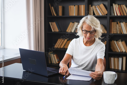 Confident stylish middle aged senior woman using laptop at workplace. Older mature lady businesswoman sitting at office table with laptop and paper document. Boss leader teacher professional worker