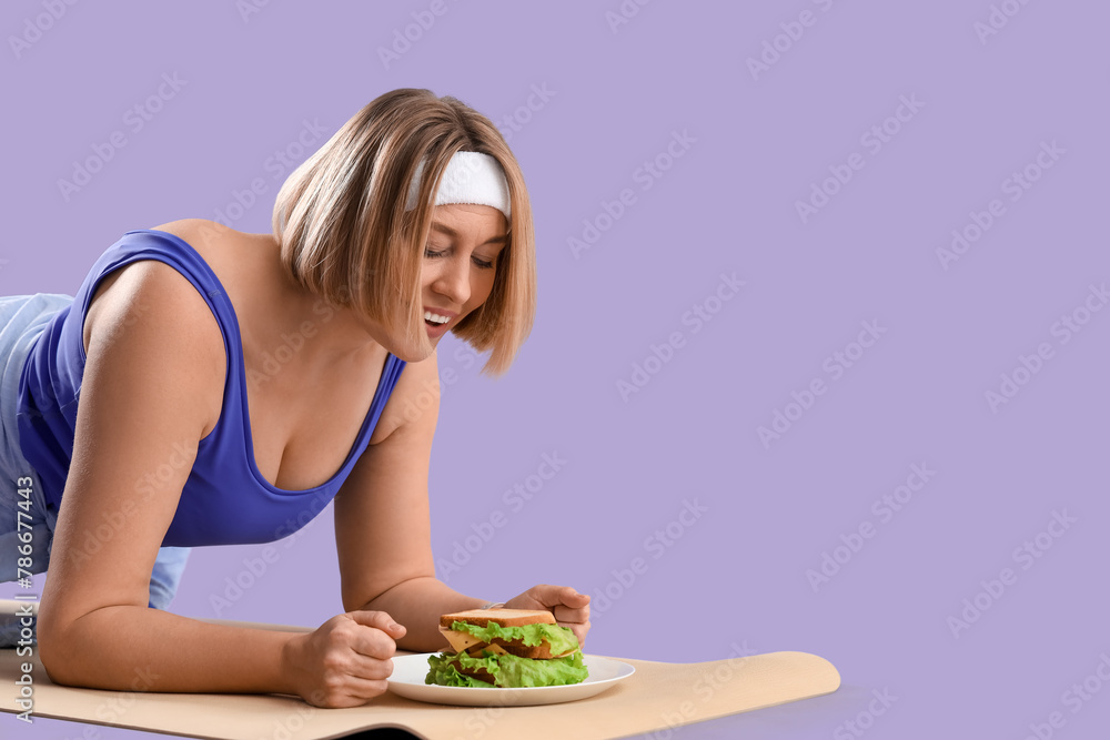 Beautiful sporty woman with sandwich training on lilac background. Overeating concept Beautiful sporty woman with sandwich training on lilac background. Overeating concept