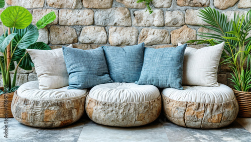 Cozy indoor setting with round cushioned seats textured stone wall, with cushions and green plants. Polished grey tiles natural light unseen source.