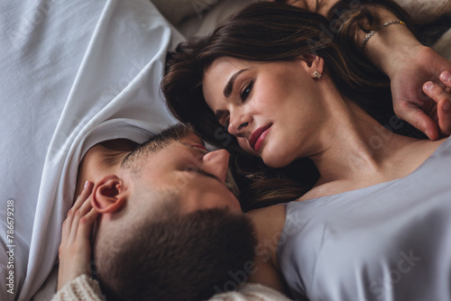 Beautiful happy loving young smiling couple relaxing in bed, looking at each other. Cozy home atmosphere, tenderness, closeness. Embracing, kissing. Lazy weekend, slow living concept