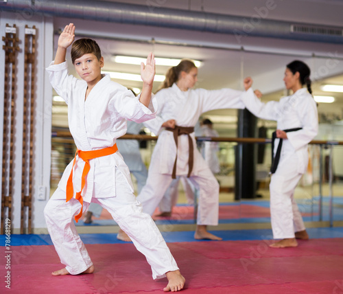Young boy in white kimono standing in fighting stance in gym. Girl sparring with trainer in background..