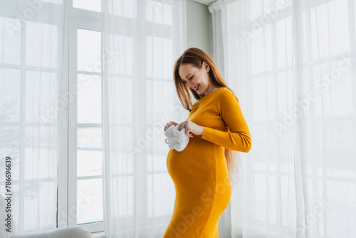Pregnancy motherhood people expectation future. Pregnant woman with big belly holding newborn baby booties smiling at home. Young mom enjoying pregnancy. Maternity tenderness parenthood new life