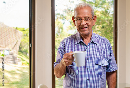 Portrait of an elderly man drinking coffee in front of a window where you can see the backyard of her house.