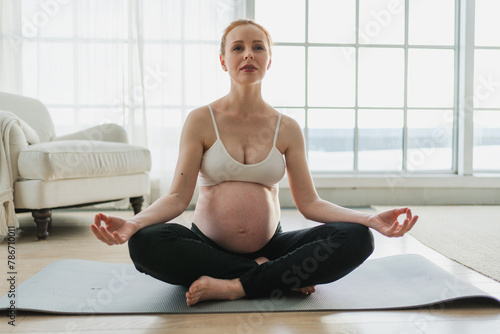 Fototapeta Naklejka Na Ścianę i Meble -  Pregnancy prenatal breathing. Pregnant woman with big belly sitting in lotus pose on yoga matt at home. Pregnant girl practicing yoga mindfulness meditation. Relaxation self care during pregnancy