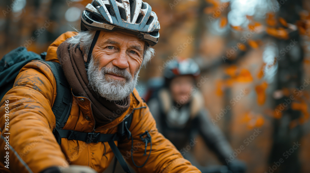 In the image, there is an elderly man wearing a helmet and a yellow jacket. He appears to be outdoors, possibly on a biking trail, with autumn foliage in the background. In the image, there is an elderly man wearing a helmet and a yellow jacket. He appears to be outdoors, possibly on a biking trail, with autumn foliage in the background.