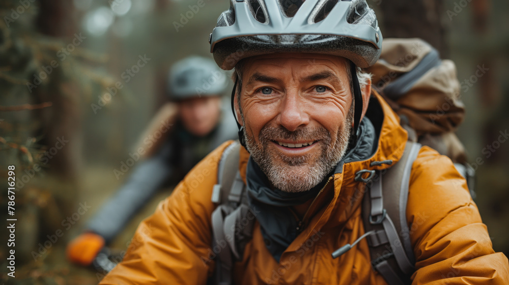 A close-up of a smiling middle-aged man wearing a cycling helmet and a yellow jacket, with a blurred background of trees. A close-up of a smiling middle-aged man wearing a cycling helmet and a yellow jacket, with a blurred background of trees.