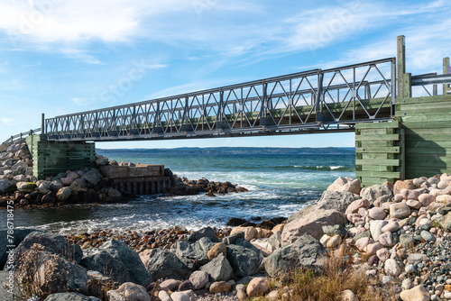 A green pedestrian wooden deck with metal railings spanning over a river entering the cold blue Atlantic Ocean. The wooden deck is made from pressure treated boards. The adjoining coastline is rocky.