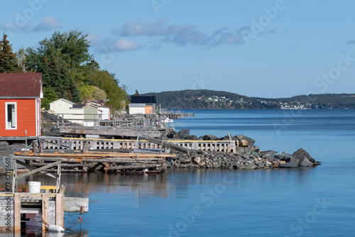 The shoreline of a small fishing village with storage sheds, wooden piers, and floating docks jutting into the calm blue ocean. The wooden buildings are for storing cod fishing supplies. 