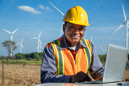 Male elderly engineer wearing a safety vest, uniform and yellow helmet using laptop in a wind farm area.