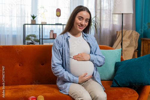 Portrait of smiling future mother sitting on couch touching pregnant belly in living room at home. Happy pregnant Caucasian lady relaxing on couch looking at camera sits near soft toy in apartment.