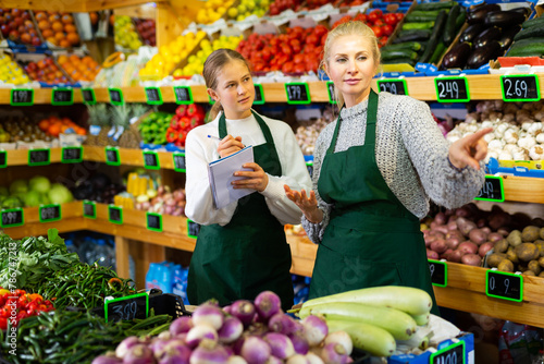 Saleswoman instructing her daughter before starting work in family store of fruits and vegetables, girl carefully writing down instructions of mother