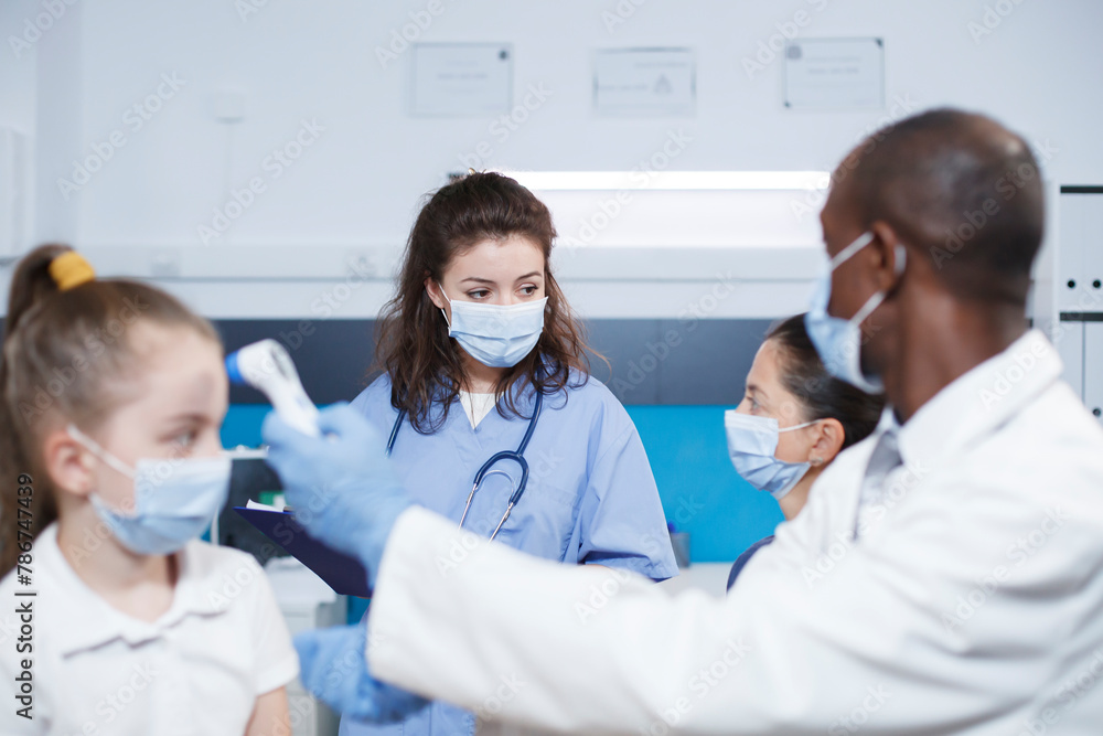 In a clinic, black doctor examines female child and communicates with the mother about health. Face mask worn, thermometer used where multiracial medical staff emphasize healthcare and medicine. In a clinic, black doctor examines female child and communicates with the mother about health. Face mask worn, thermometer used where multiracial medical staff emphasize healthcare and medicine.