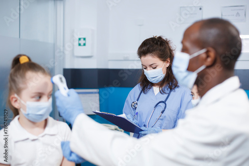 Woman in blue scrubs writing notes while black man in a lab coat checks temperature of caucasian child. Image shows male doctor examining patient with assistance from nurse. photo