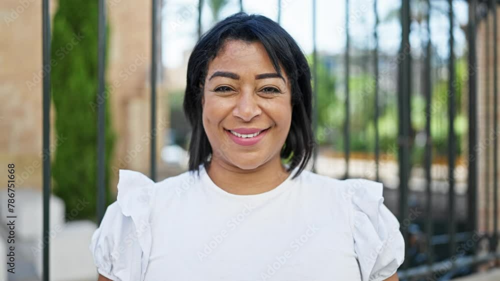 Smiling hispanic woman in white shirt standing outdoors near metal gates in a sunny urban park setting. Smiling hispanic woman in white shirt standing outdoors near metal gates in a sunny urban park setting.