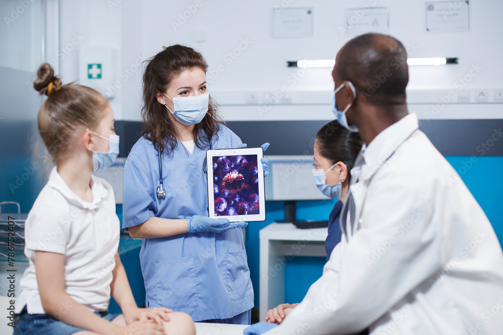 Woman wearing blue scrubs holding a digital tablet presenting a virus cell picture in front of a girl and her mother, and the african american male physician. Expert treatment and education provided. Woman wearing blue scrubs holding a digital tablet presenting a virus cell picture in front of a girl and her mother, and the african american male physician. Expert treatment and education provided.