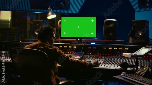 African american music producer working with greenscreen on pc, using mixing console to do mix and master in control room. Audio technician recording tracks with knobs and switchers. Camera B.
