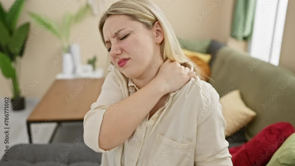 A young woman experiencing neck pain while sitting on a couch indoors A young woman experiencing neck pain while sitting on a couch indoors