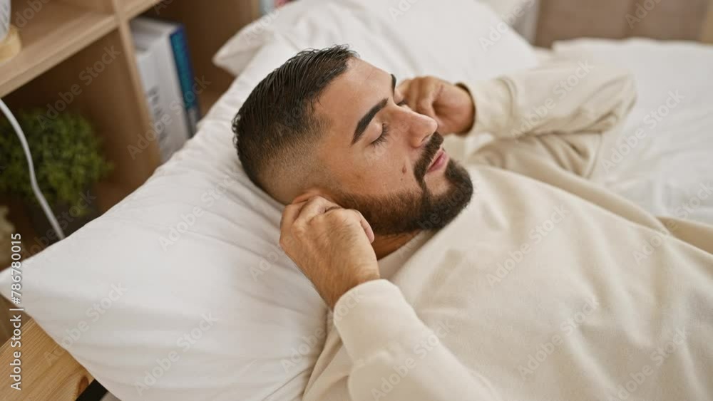 A relaxed young man with a beard enjoys music on wireless earphones while lying in bed at his cozy interior home. A relaxed young man with a beard enjoys music on wireless earphones while lying in bed at his cozy interior home.