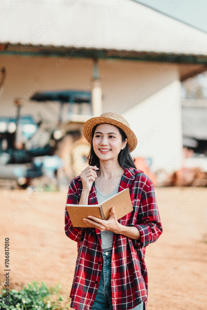 Obraz premium Smiling farmer woman stands holding a notebook, dressed in a straw hat and plaid shirt, with a tractor in the background.