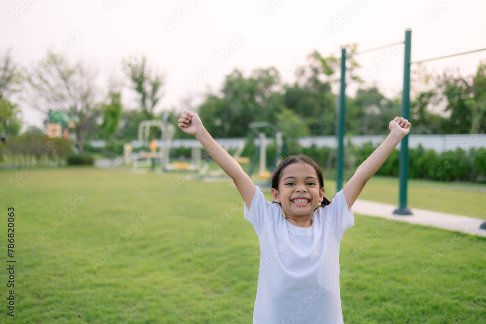 A young girl is standing in a park, smiling and raising her arms in the air A young girl is standing in a park, smiling and raising her arms in the air