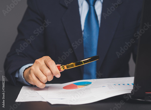 Businessman holding a magnifying glass and looking at reading documents while sitting at a desk.
