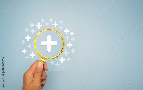 Hand holding a magnifying glass against a light blue background with white plus symbols.