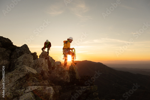 Two people are climbing a mountain, one of them wearing a yellow jacket. The sun is setting in the background, creating a beautiful and serene atmosphere