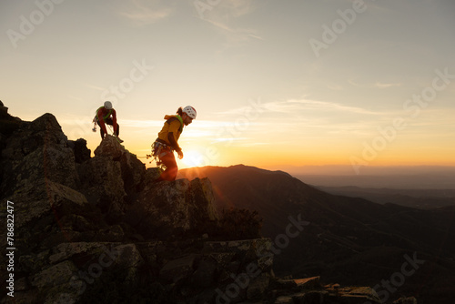 Two people are climbing a mountain and the sun is setting in the background. Scene is adventurous and exciting