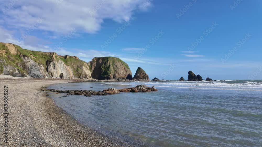 Crystal clear water gentle waves on pebble beach with sea cliffs and blue sky with puffy wihite clouds bright spring day Copper Coast Waterford Ireland Crystal clear water gentle waves on pebble beach with sea cliffs and blue sky with puffy wihite clouds bright spring day Copper Coast Waterford Ireland