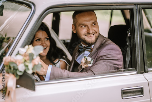 Valmiera, Latvia- July 28, 2024 - Bride and groom smiling inside a classic car, groom at the wheel, bride with a bouquet, both looking joyfully at each other.